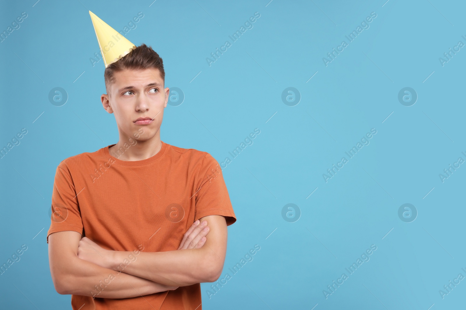 Photo of Young man in party hat with crossed arms on light blue background. Space for text