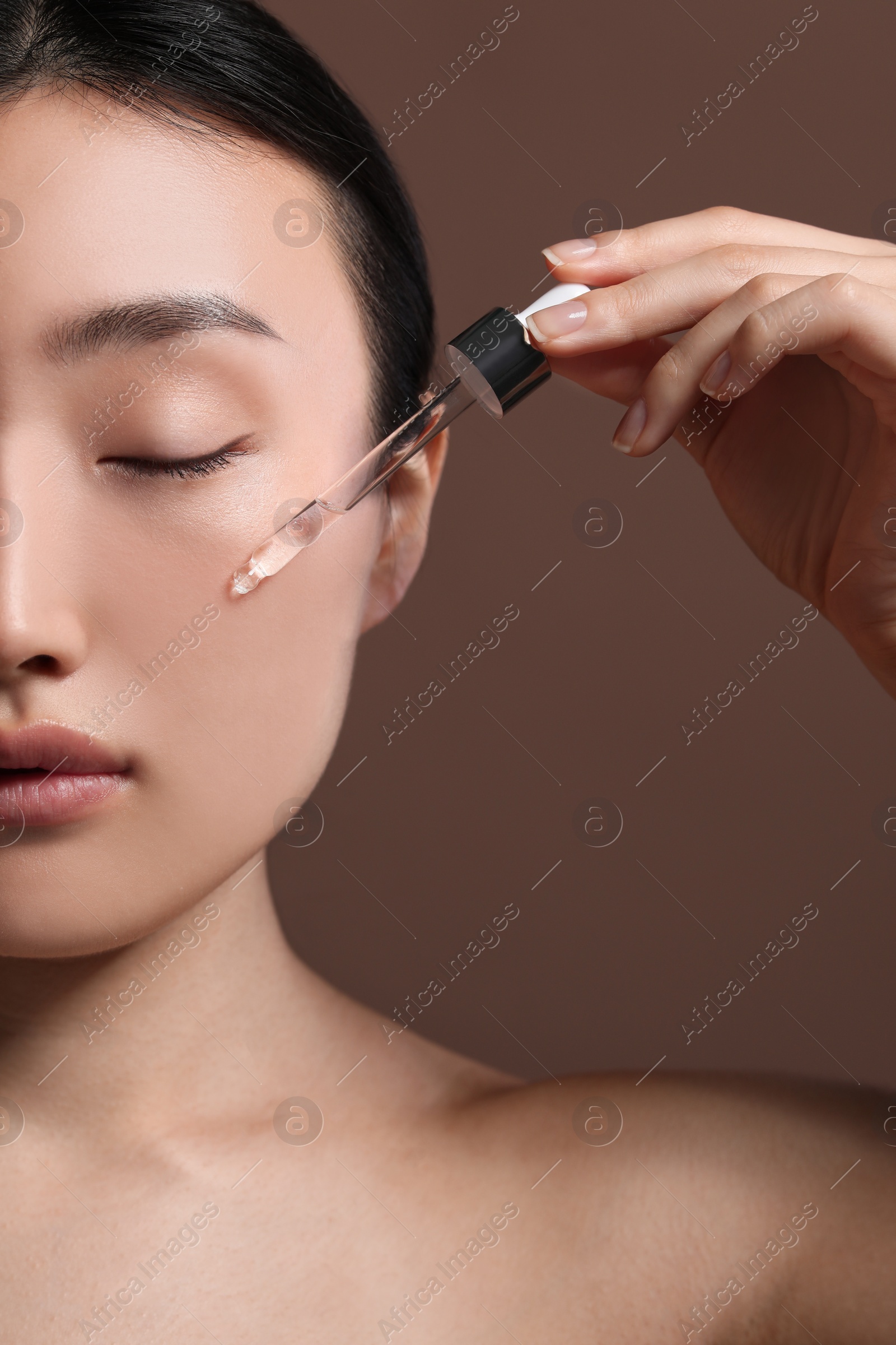 Photo of Beautiful young woman applying cosmetic serum onto her face on brown background, closeup