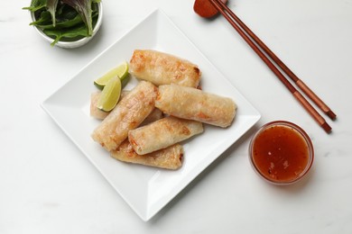 Photo of Tasty fried spring rolls, lime, sauce and lettuce served on white marble table, flat lay