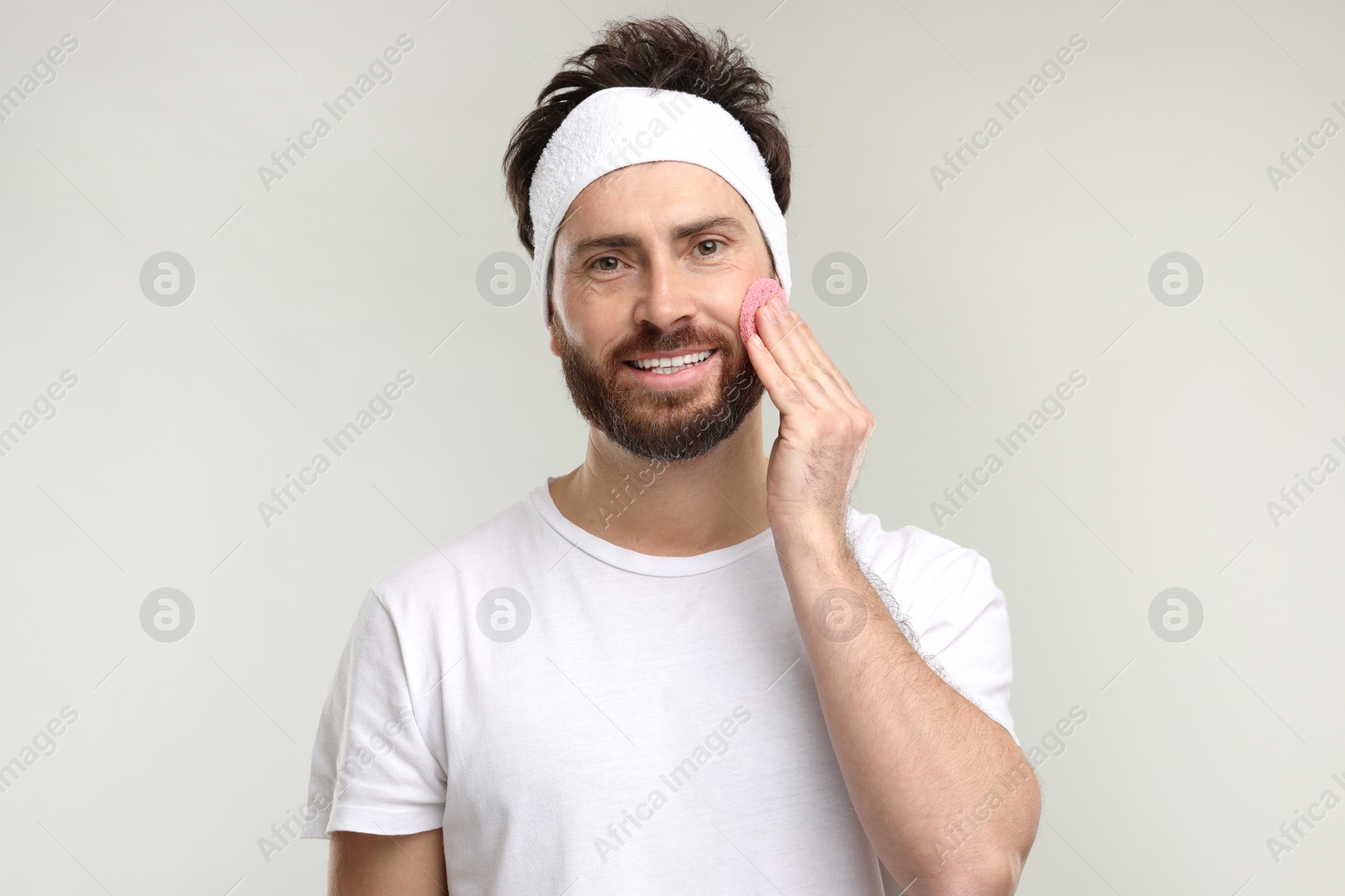 Photo of Man with headband washing his face using sponge on light grey background