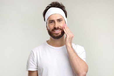 Man with headband washing his face using sponge on light grey background