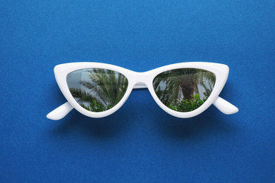 Stylish sunglasses with reflection of palm trees on blue background, top view 