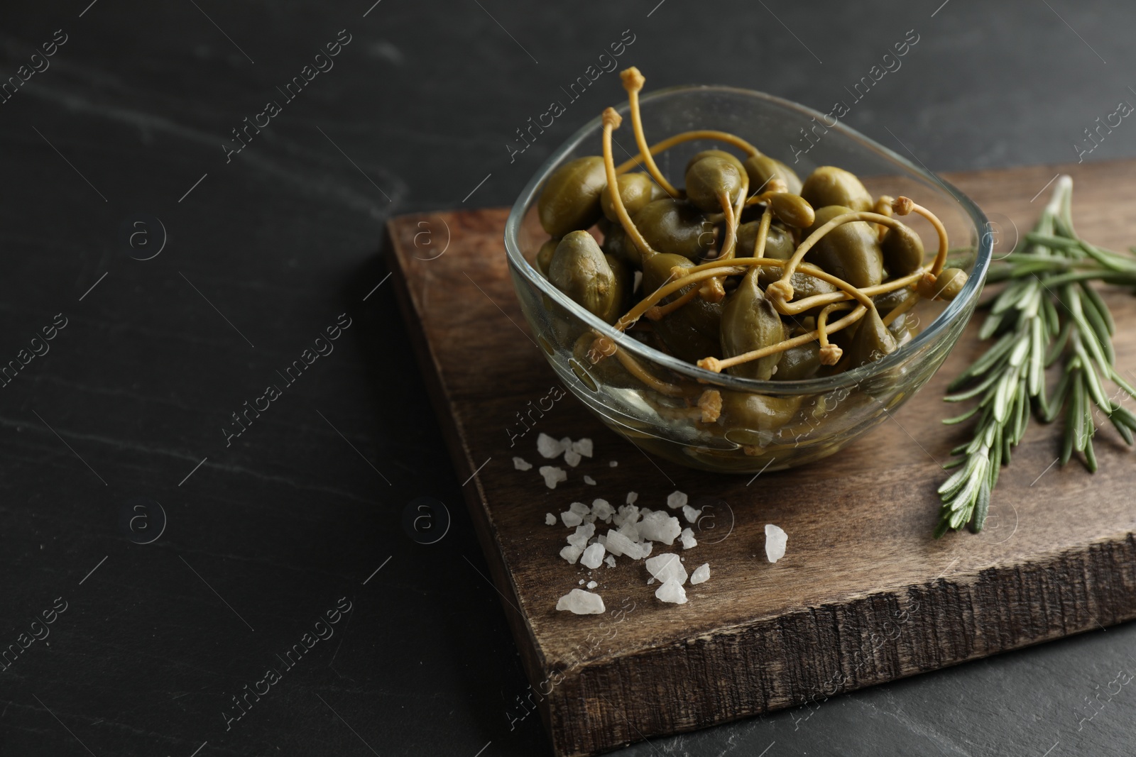 Photo of Tasty capers in glass bowl, salt and rosemary on black table, space for text