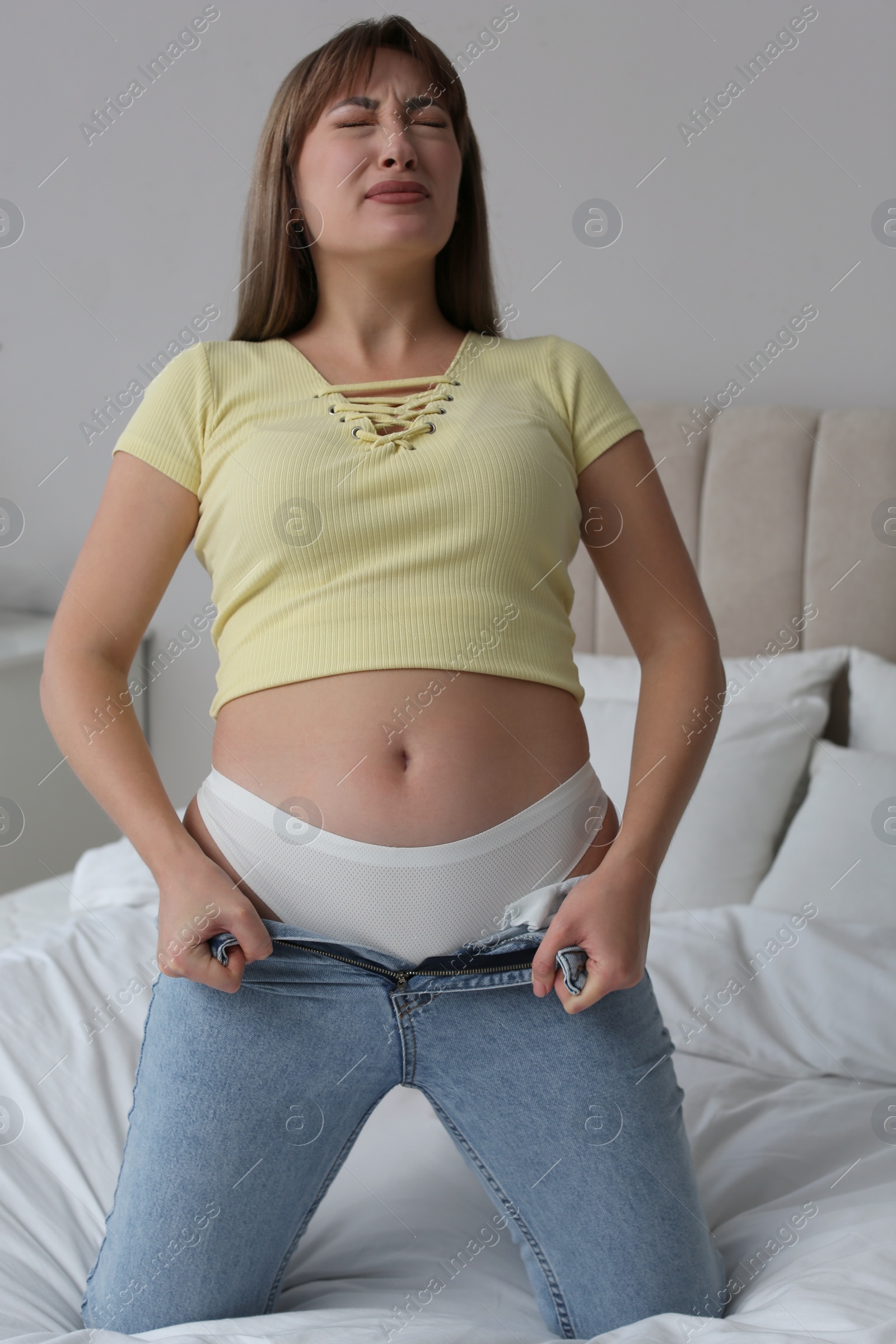 Photo of Young woman struggling to squeeze into tight jeans on bed at home