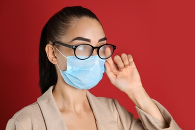 Photo of Woman wiping foggy glasses caused by wearing medical mask on red background