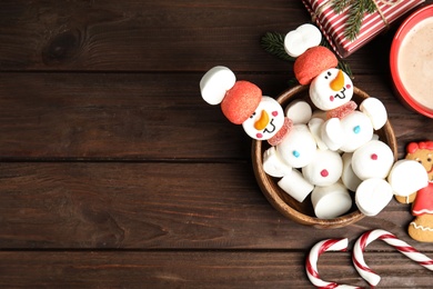 Photo of Flat lay composition with funny snowmen made of marshmallows in bowl on wooden table, space for text