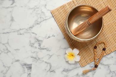 Golden singing bowl with mallet, beads and flower on white marble table, flat lay. Space for text