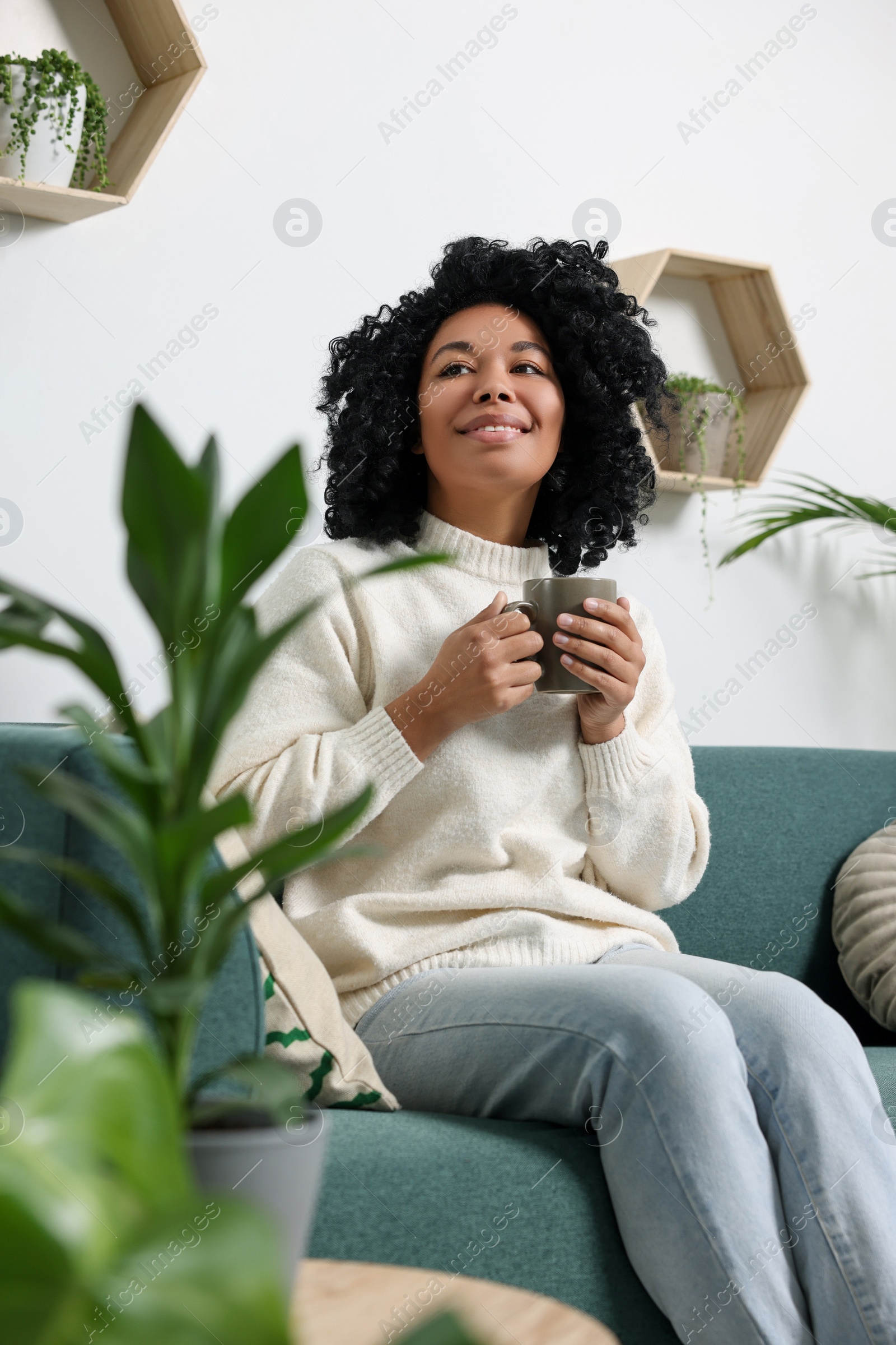 Photo of Relaxing atmosphere. Happy woman with cup of hot drink on sofa near beautiful houseplants in room