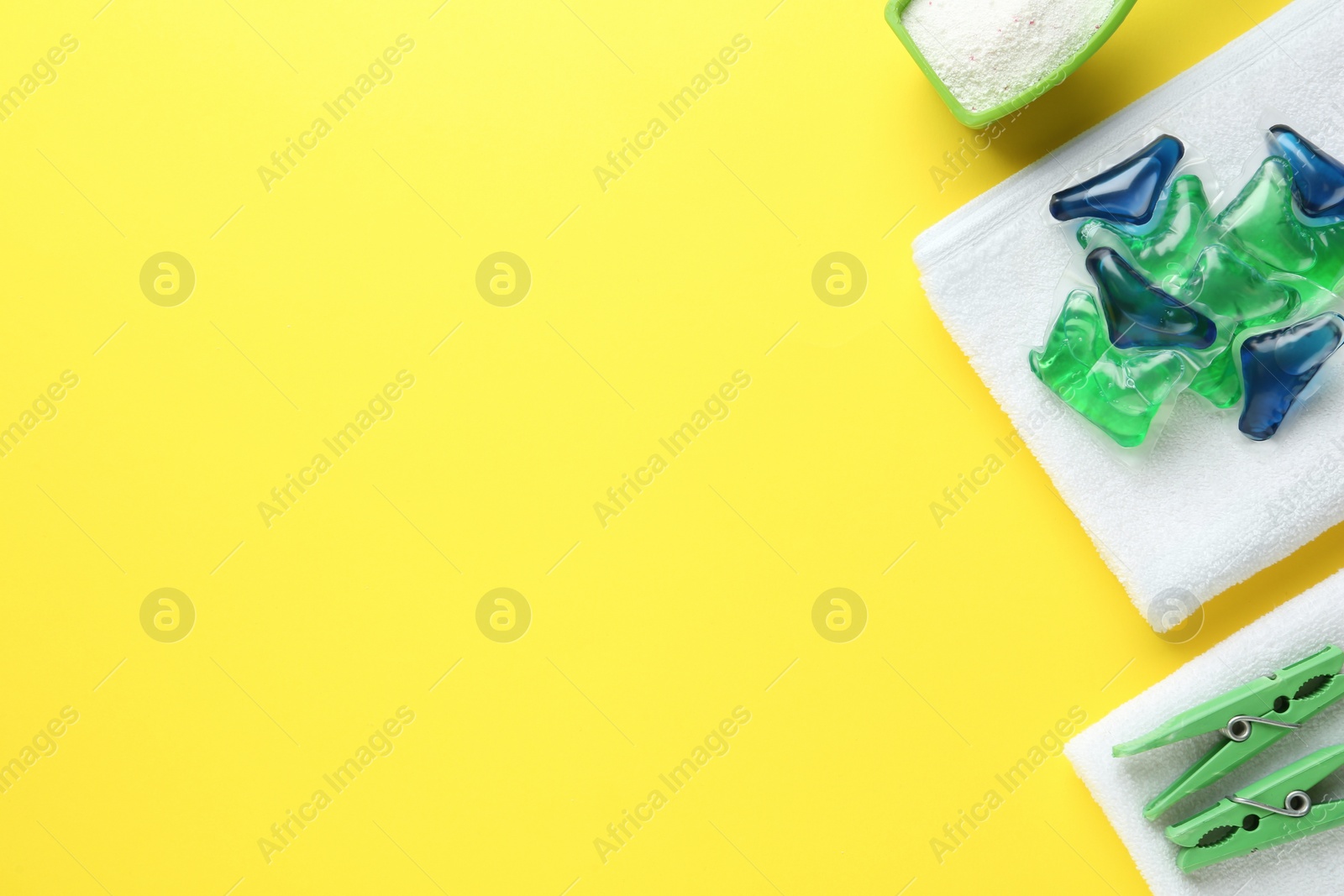 Photo of Laundry capsules, washing powder, towels and clothespins on yellow background, flat lay. Space for text