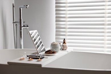 Photo of White wooden tray with tablet, spa products and burning candles on bathtub in bathroom