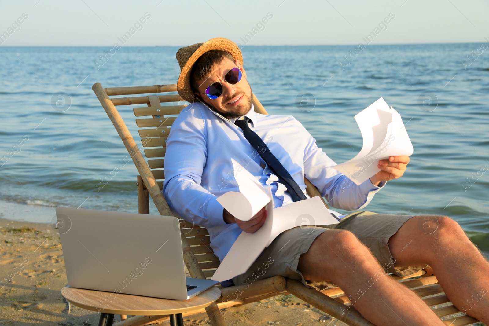 Photo of Busy man with papers talking by mobile phone on beach. Business trip