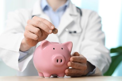 Photo of Doctor putting coin into piggy bank at table, closeup