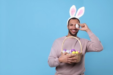Photo of Happy African American man in bunny ears headband covering eye with Easter egg on light blue background, space for text