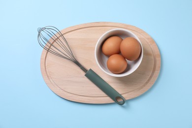 Photo of Metal whisk and raw eggs on light blue background, top view