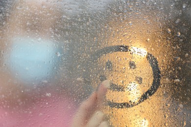 Photo of Woman drawing happy face on foggy window at rainy weather, closeup
