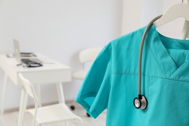 Turquoise medical uniform and stethoscope on rack in clinic, closeup. Space for text