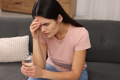 Sad woman with glass of water suffering from headache on sofa indoors