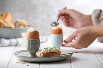 Woman eating breakfast with fresh soft boiled egg at white wooden table, focus on plate