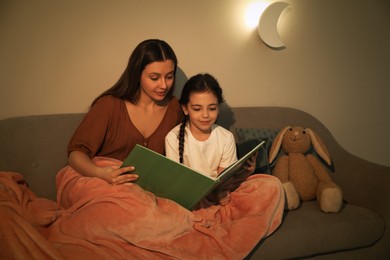 Photo of Little girl with mother reading book in living room lit by night lamp