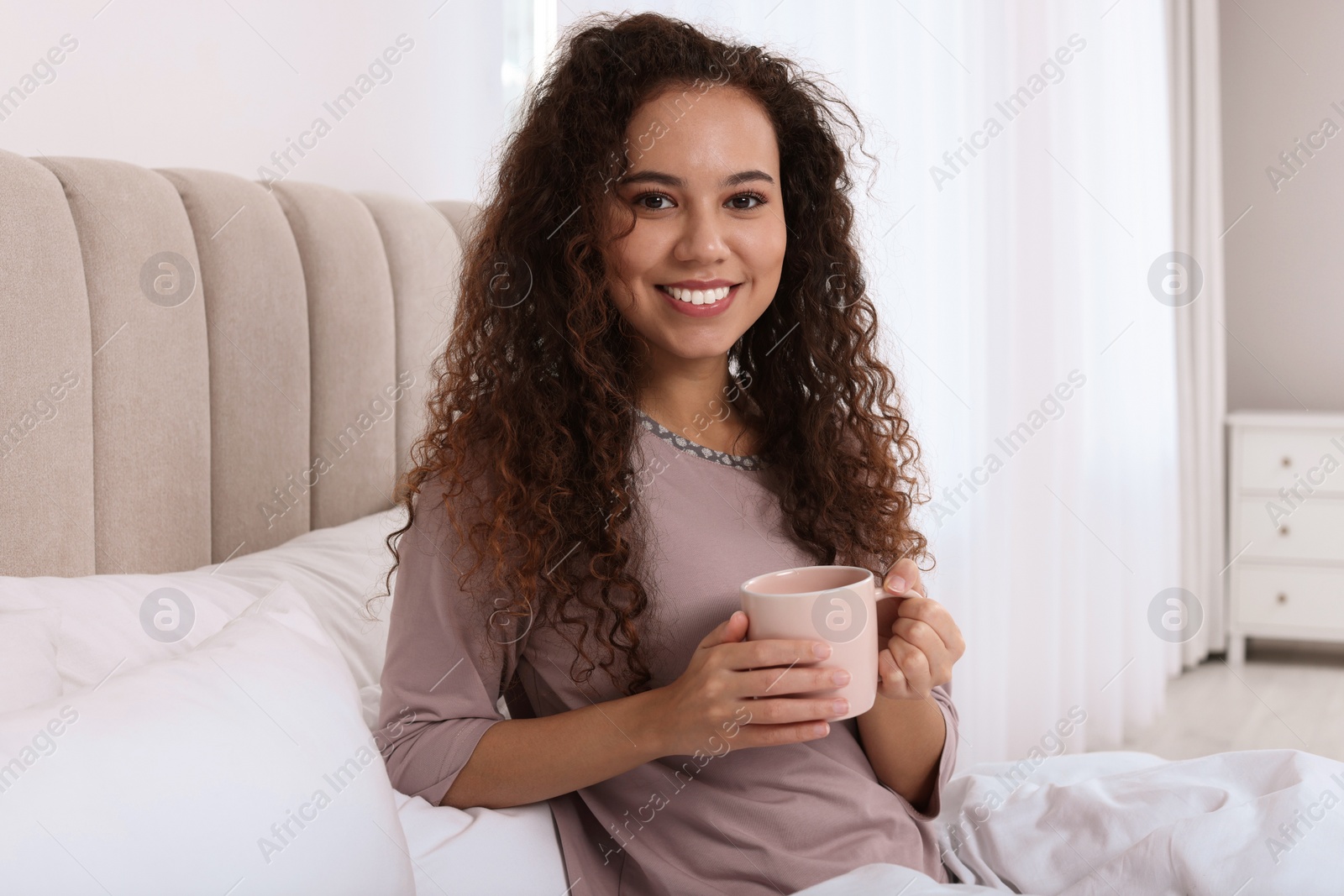 Photo of Happy African American woman with cup of drink in bed at home