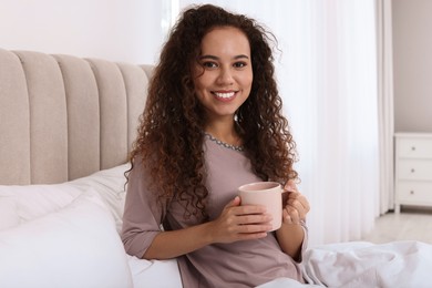 Happy African American woman with cup of drink in bed at home