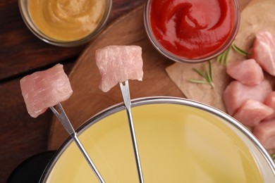 Fondue pot, forks with pieces of raw meat and sauces on wooden table, flat lay