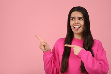 Photo of Happy young woman showing her tongue and pointing on pink background. Space for text