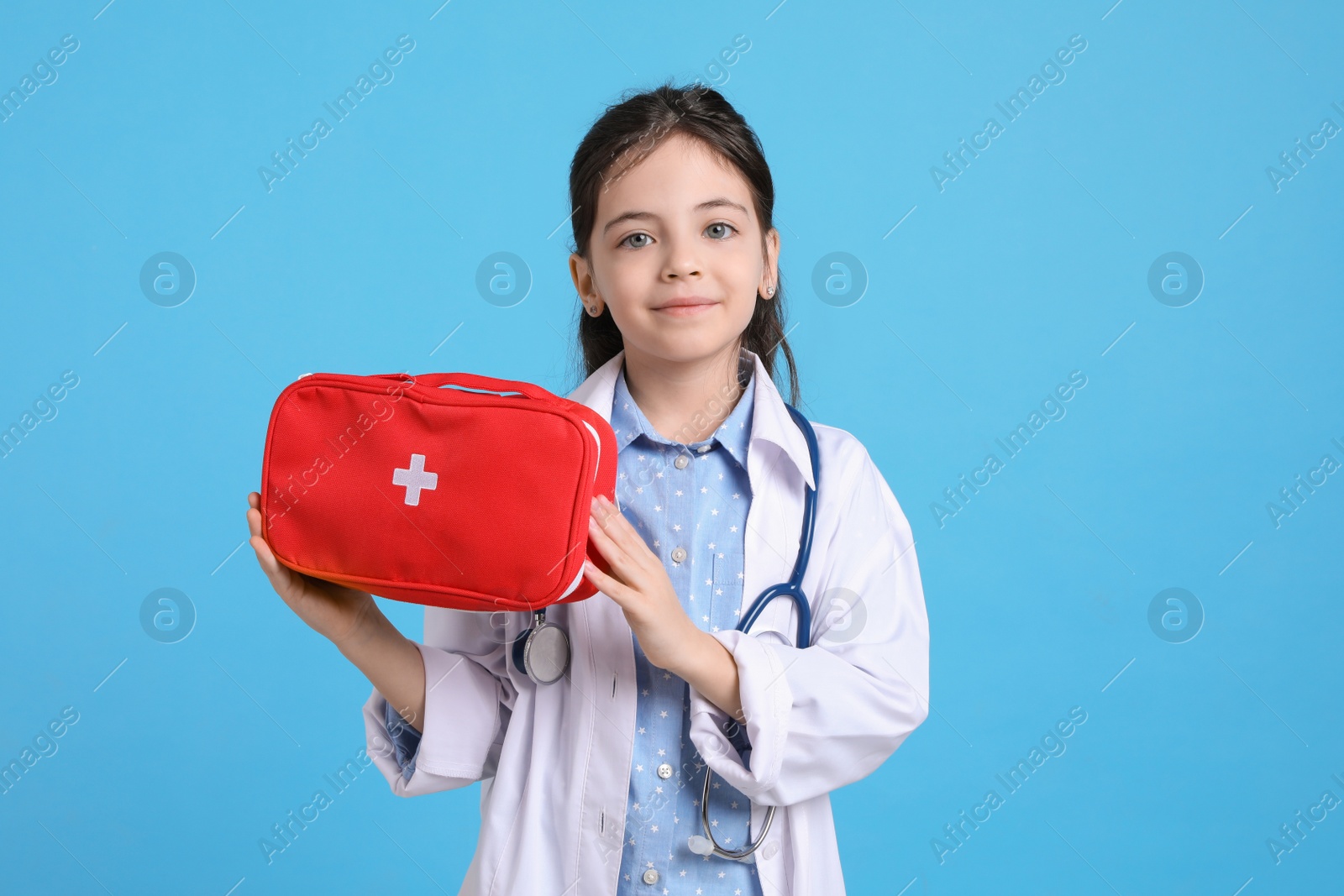 Photo of Little girl with first aid bag and stethoscope dressed as doctor on light blue background. Pediatrician practice