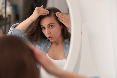 Photo of Emotional woman with hair loss problem looking in mirror indoors