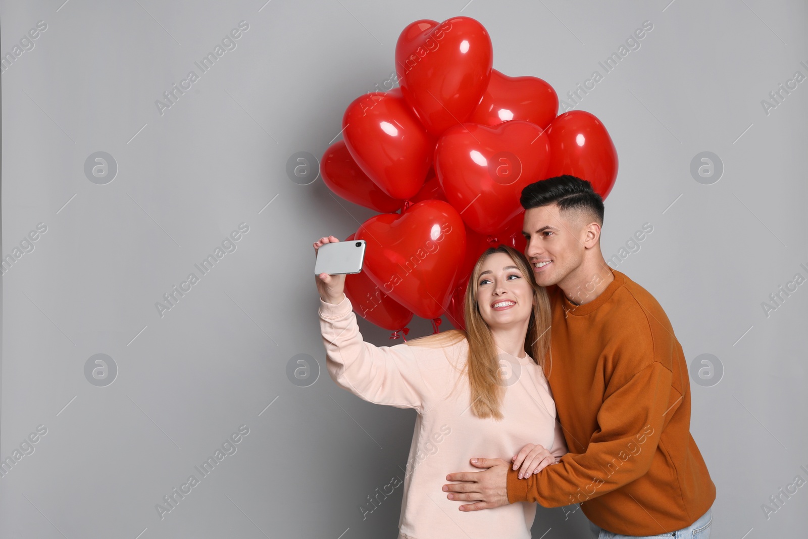 Photo of Lovely couple with heart shaped balloons taking selfie on grey background. Valentine's day celebration