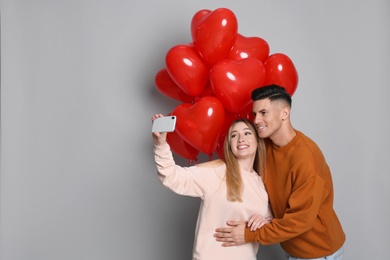 Photo of Lovely couple with heart shaped balloons taking selfie on grey background. Valentine's day celebration