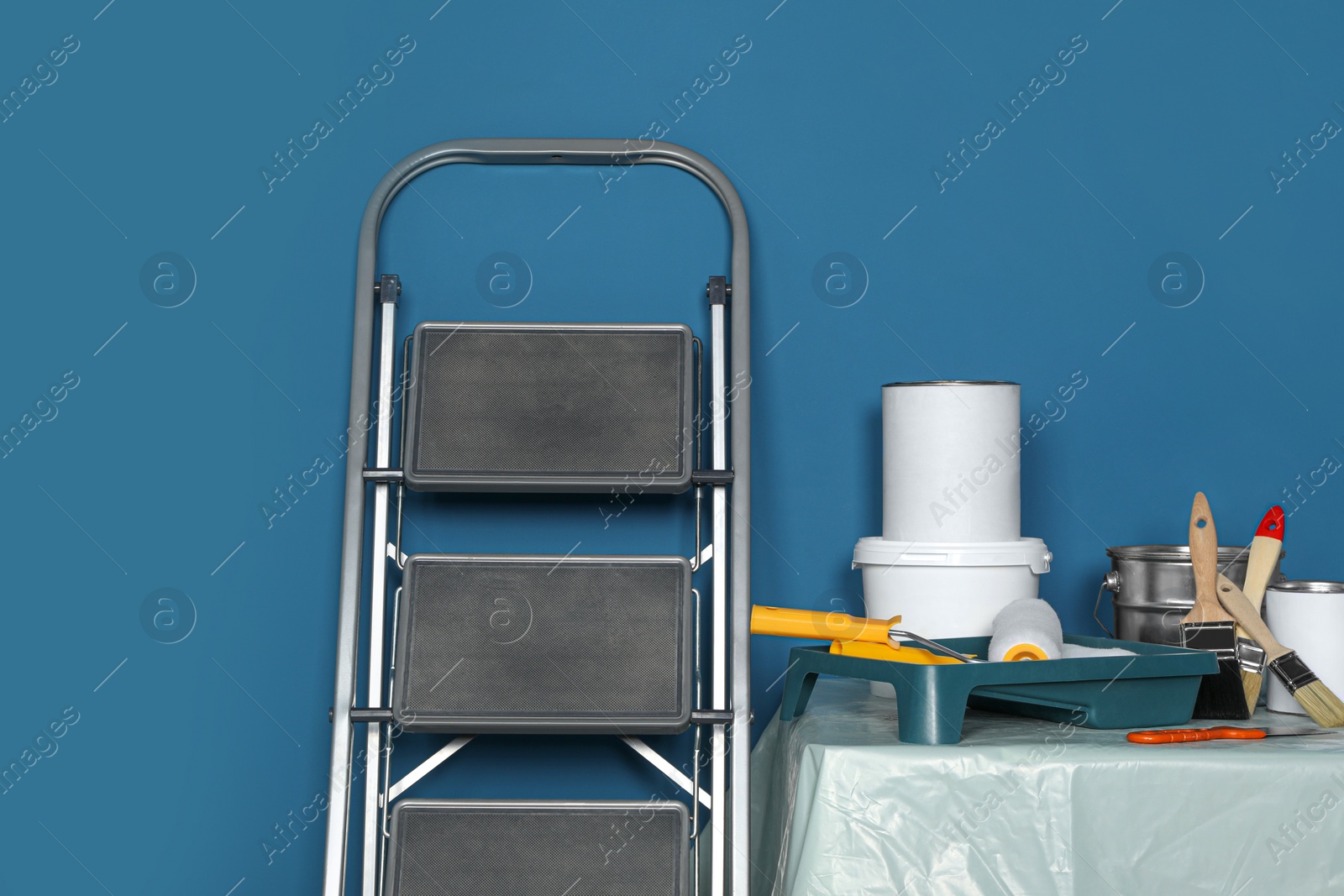 Photo of Metallic folding ladder and painting tools on table near blue wall