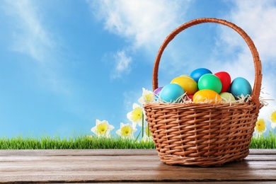 Image of Colorful Easter eggs in wicker basket on wooden table outdoors, space for text 