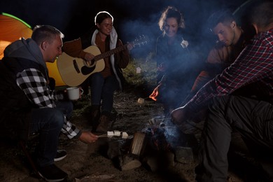 Group of friends roasting marshmallows on bonfire at camping site in evening