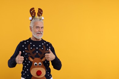 Photo of Senior man in Christmas sweater and reindeer headband showing thumbs up on orange background. Space for text
