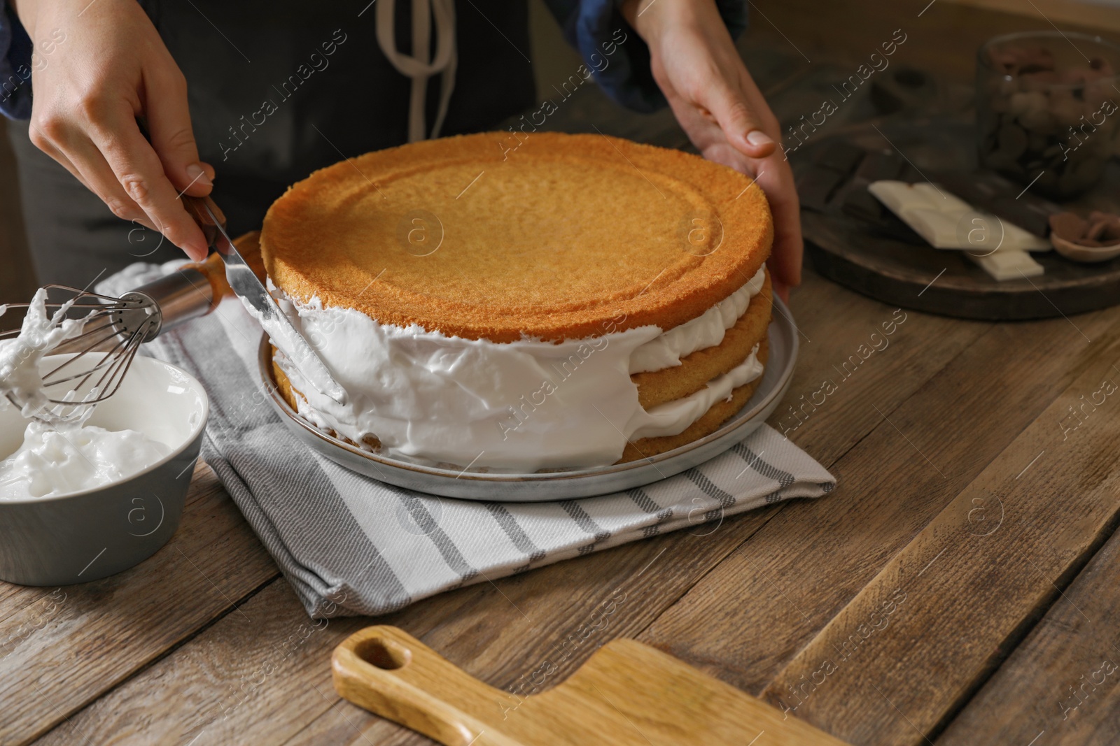 Photo of Woman smearing sides of sponge cake with cream at wooden table, closeup