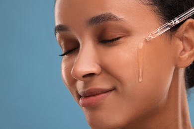 Beautiful woman applying serum onto her face on blue background, closeup