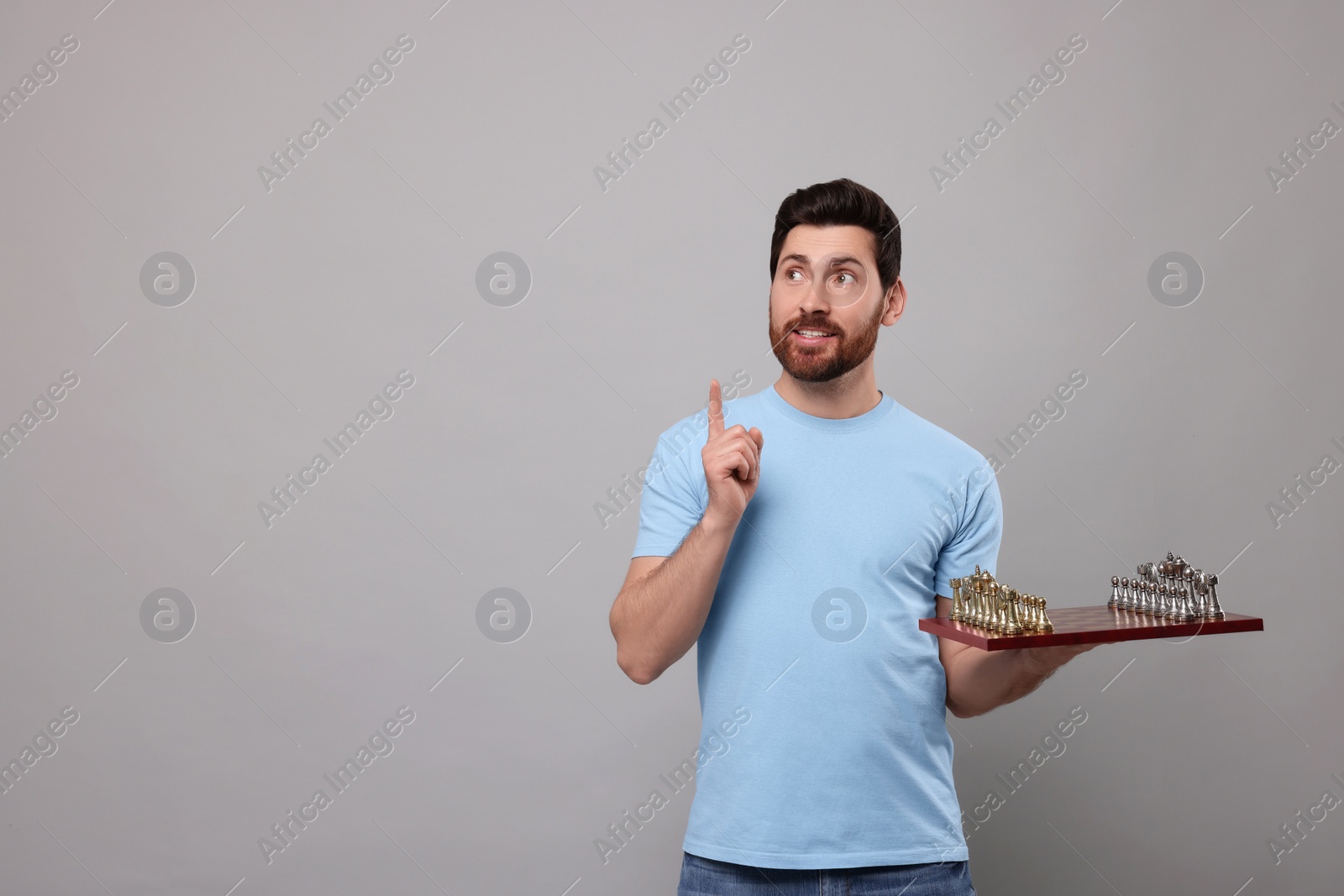 Photo of Handsome man holding chessboard with game pieces and pointing at something on light grey background. Space for text