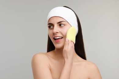 Photo of Young woman with headband washing her face using sponge on light grey background