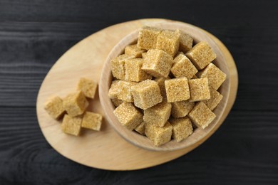 Photo of Brown sugar cubes in bowl on black wooden table, top view