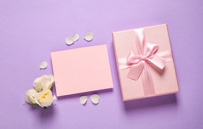 Photo of Pink gift box, blank card and beautiful flowers on violet background, flat lay. Space for text