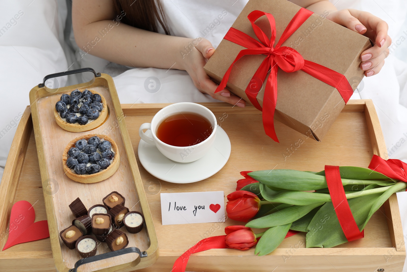 Photo of Tasty breakfast served in bed. Woman with gift box, desserts, tea, flowers and I Love You card at home, closeup