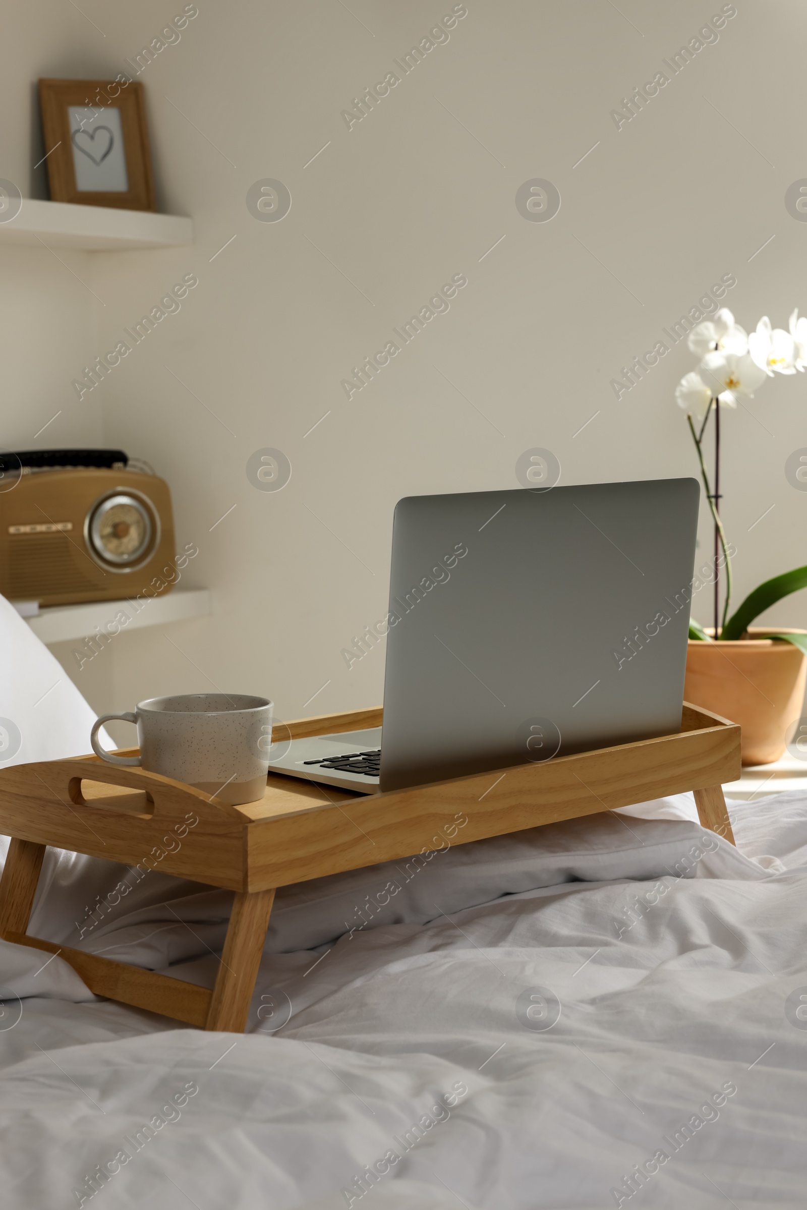 Photo of Wooden tray with modern laptop and cup of aromatic drink on bed indoors