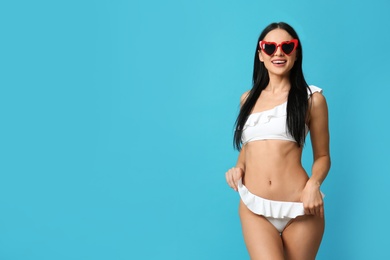 Beautiful young woman in white bikini with sunglasses on light blue background. Space for text