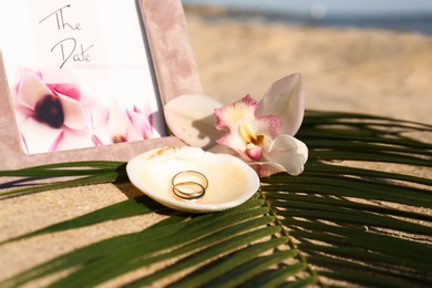 Photo of Shell with gold rings, wedding invitation and beautiful flower on sandy beach