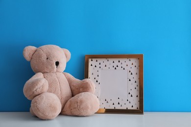 Empty photo frame and teddy bear on white table near light blue wall. Space for design