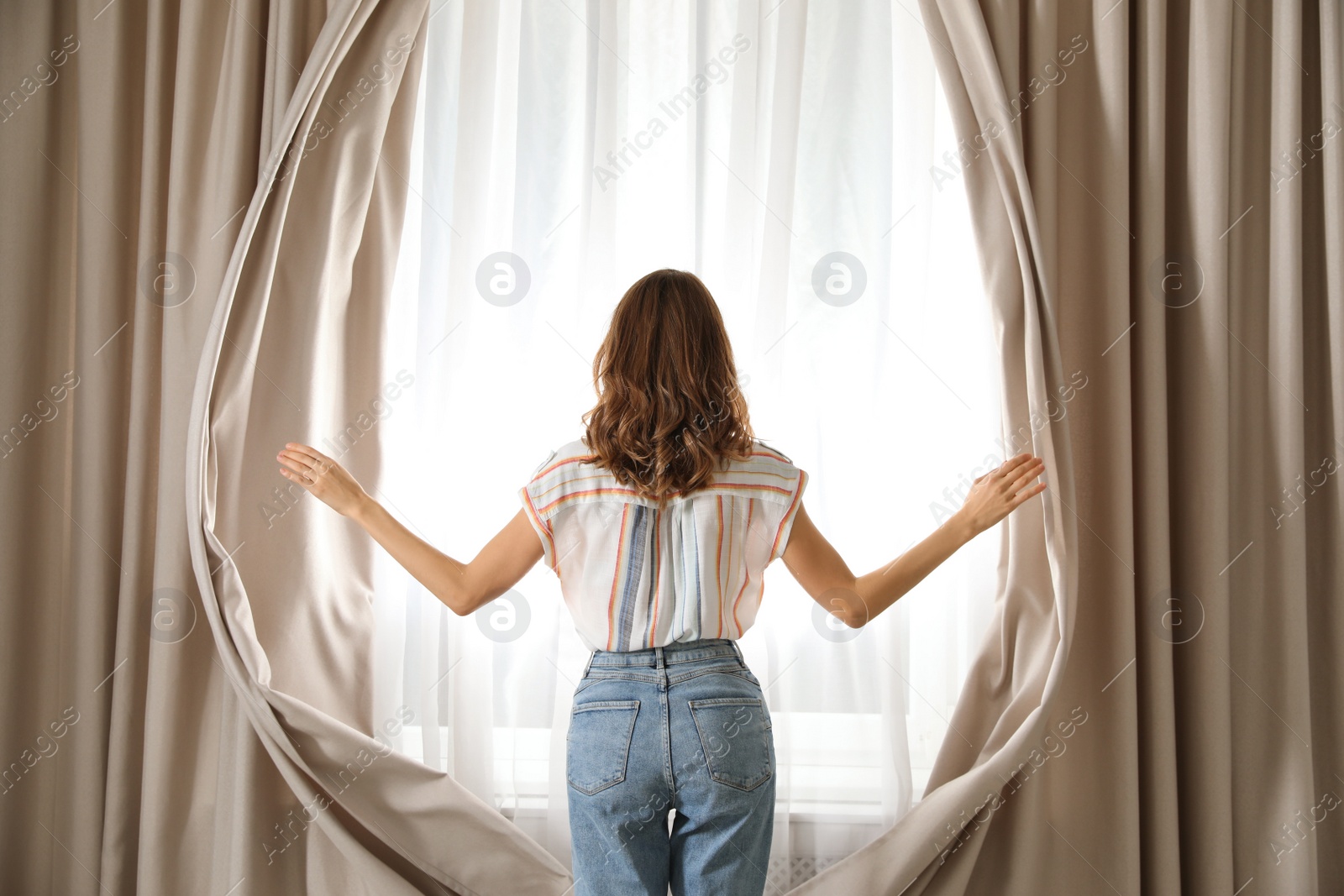 Photo of Woman opening window curtains at home in morning, back view