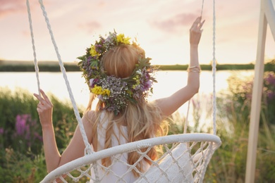 Photo of Young woman wearing wreath made of beautiful flowers on swing chair outdoors, back view