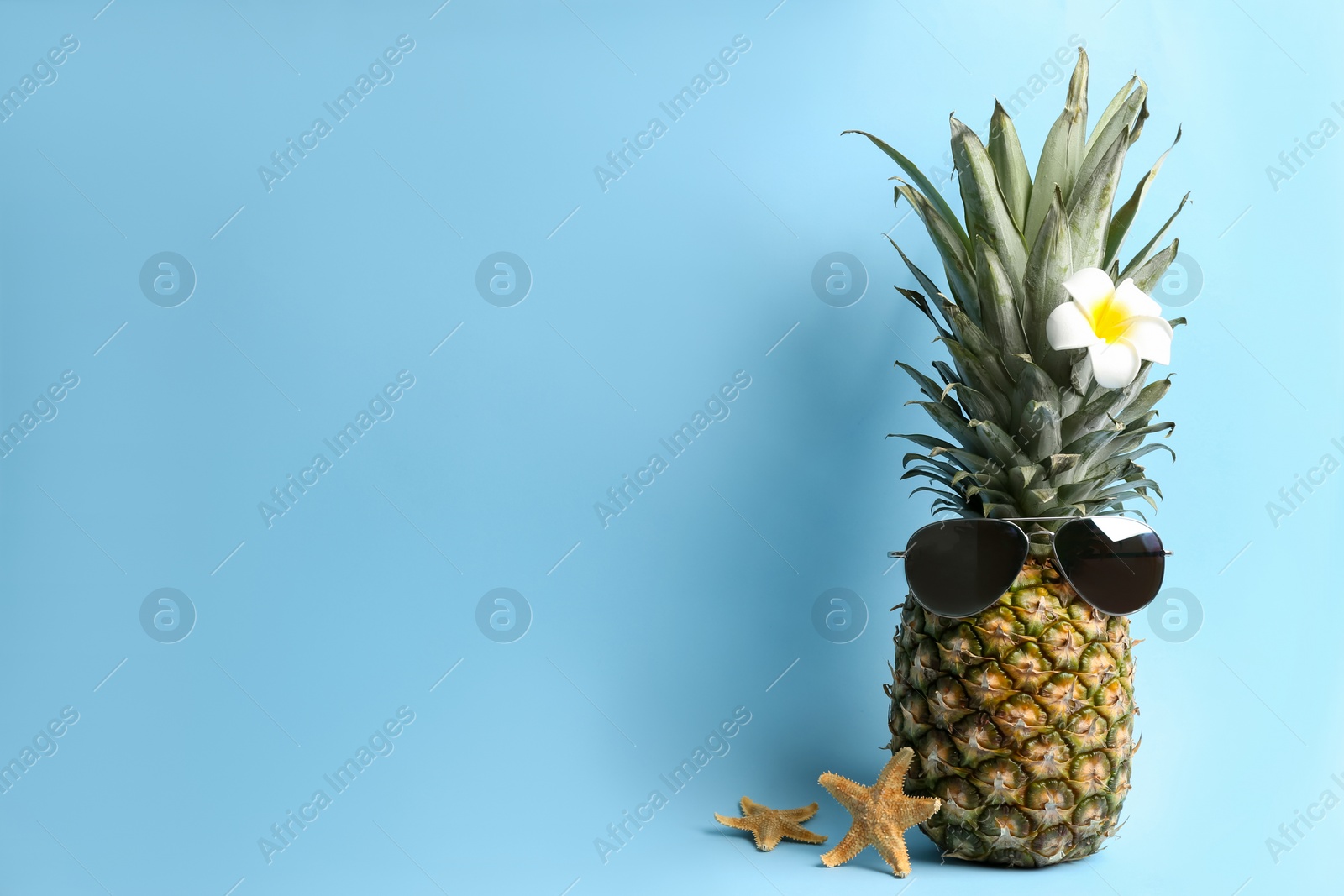 Photo of Pineapple with sunglasses, plumeria flower and starfishes on light blue background, space for text. Creative concept