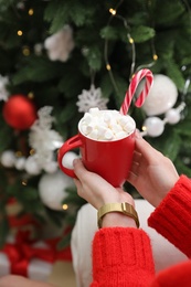 Photo of Woman with cup of delicious hot drink near Christmas tree indoors, closeup